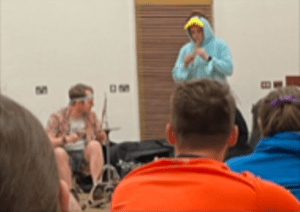 Two young people making music - one is wearing a turquoise onesie with a yellow beak-like hood