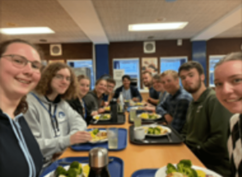 11 smiling young people sitting at a long dinner table with trays of food in front of each of them