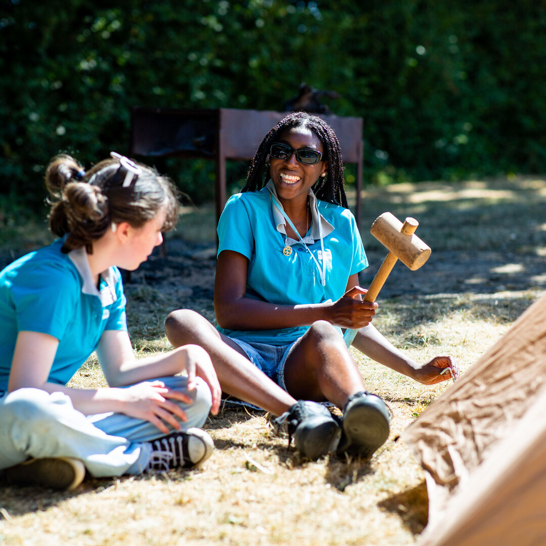 Two girls, aged between 14-18, sit outdoors, smiling while putting up a tent