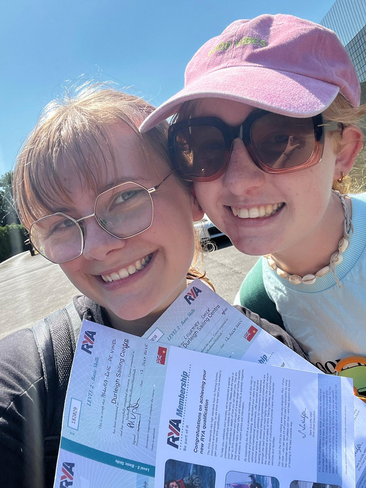 Close up photo of two young women smiling, holding up their RYA level 2 sailing certificates, with a bright blue sky in the background