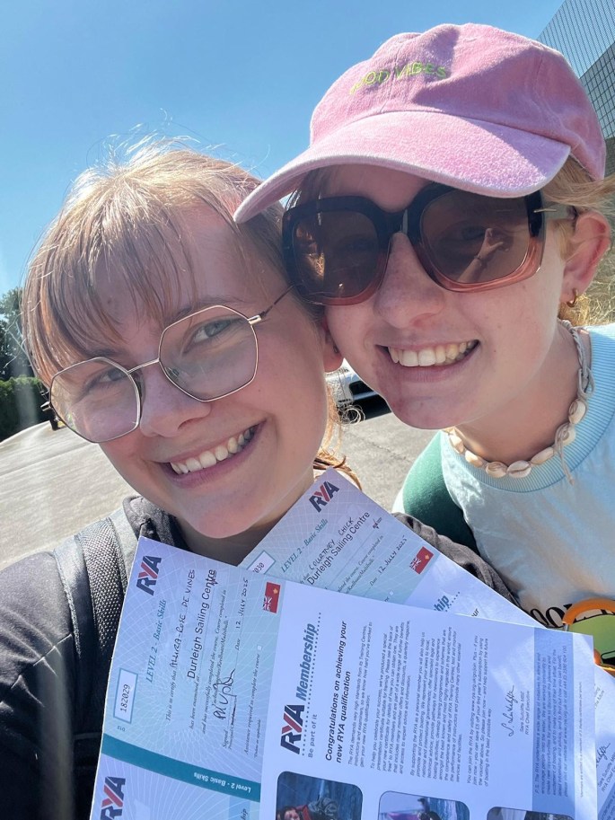 Close up photo of two young women smiling, holding up their RYA level 2 sailing certificates, with a bright blue sky in the background